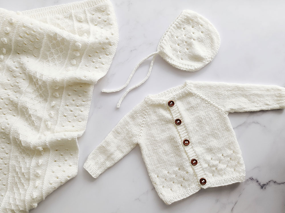 Off white baby blanket with bobbles, eyelets and lattice pattern. Off white eyelet patterned baby cardigan and off white eyelet pattern baby bonnet